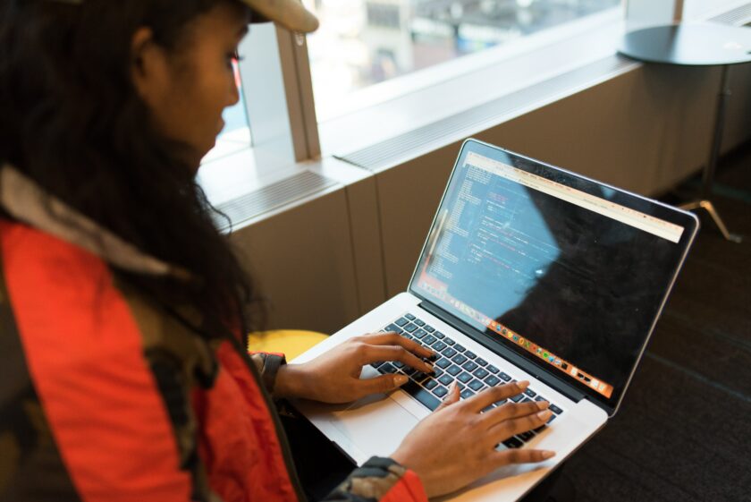 Woman wearing a hat and black and red jacket typing on her laptop.