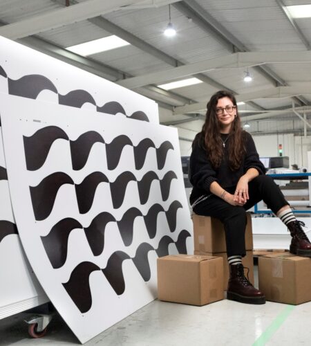 Katie McAfee, Director of FYI, sitting on cardboard boxes next to large white panels with a printed black stylized wave design. The panels are stacked on a trolley at one of FYI's warehouses.