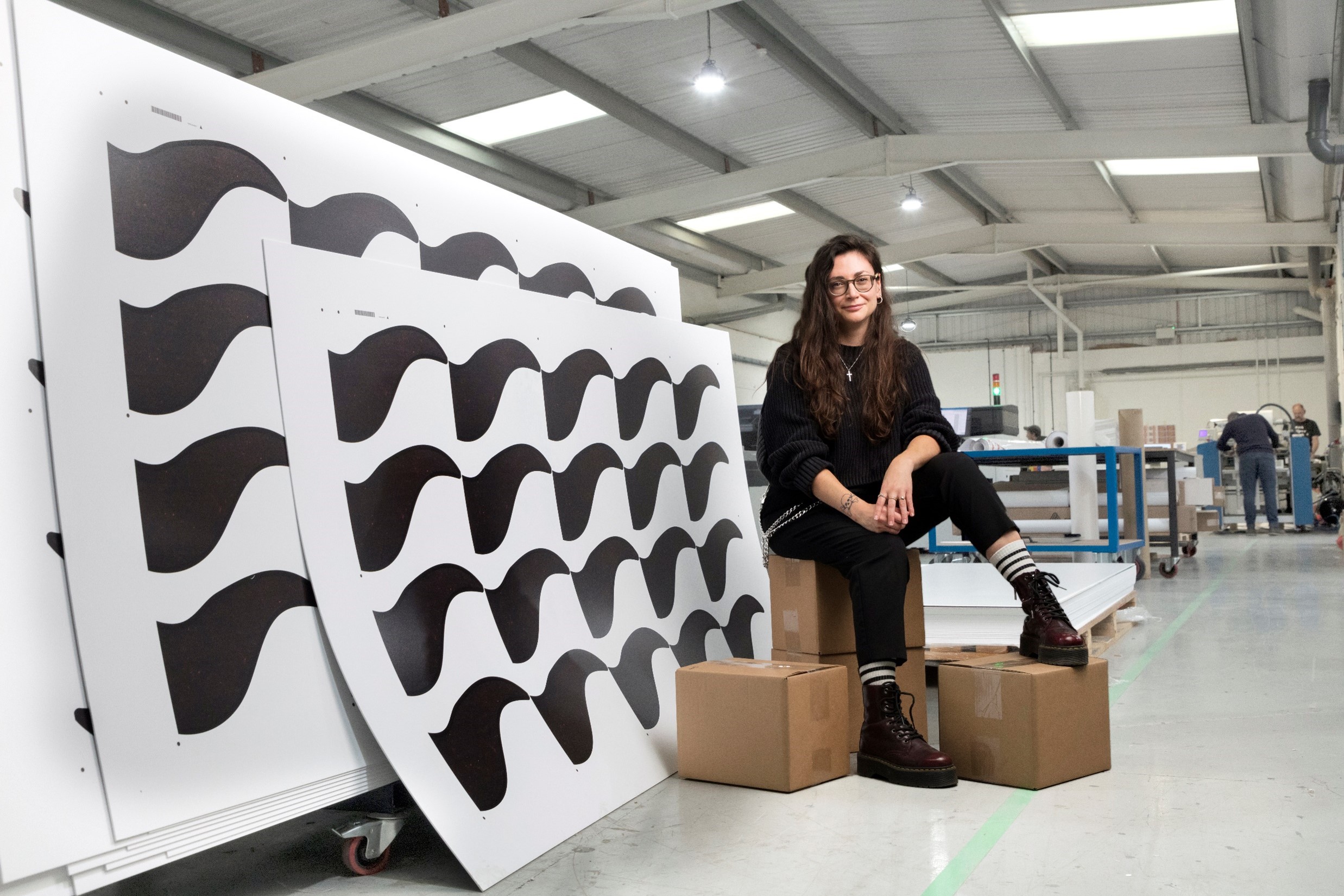 Katie McAfee, Director of FYI, sitting on cardboard boxes next to large white panels with a printed black stylized wave design. The panels are stacked on a trolley at one of FYI's warehouses.