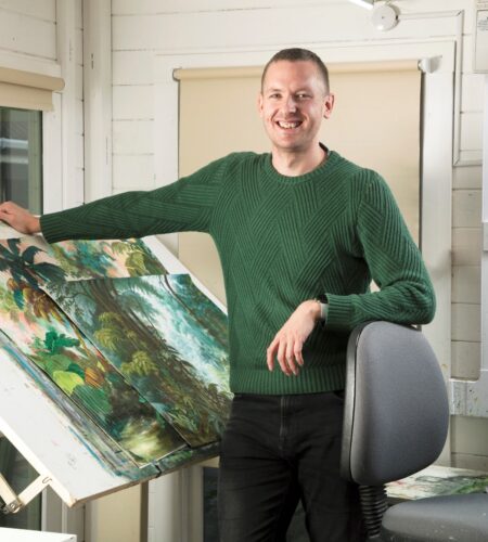 Richard Haley, MD, is wearing a green jumper and standing next to a white easel in the business's art studio. Clipped to the easel is two pieces of artwork, the first and most prominent painting depicts a tropical scene with palm trees and lush foliage.